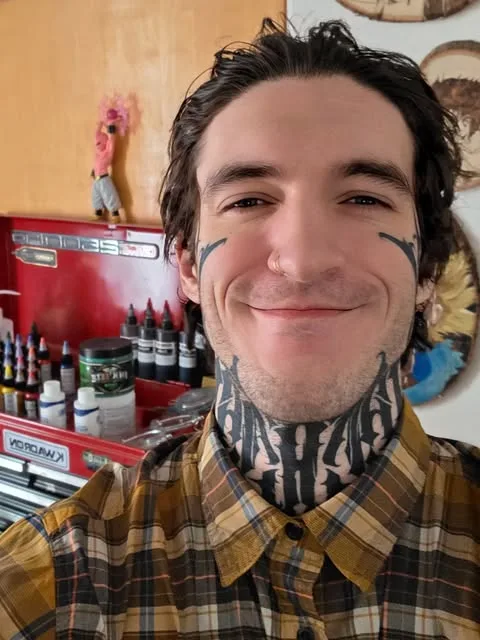 Nicholas Lally smiling at his station, with a shelf of tattoo inks behind him